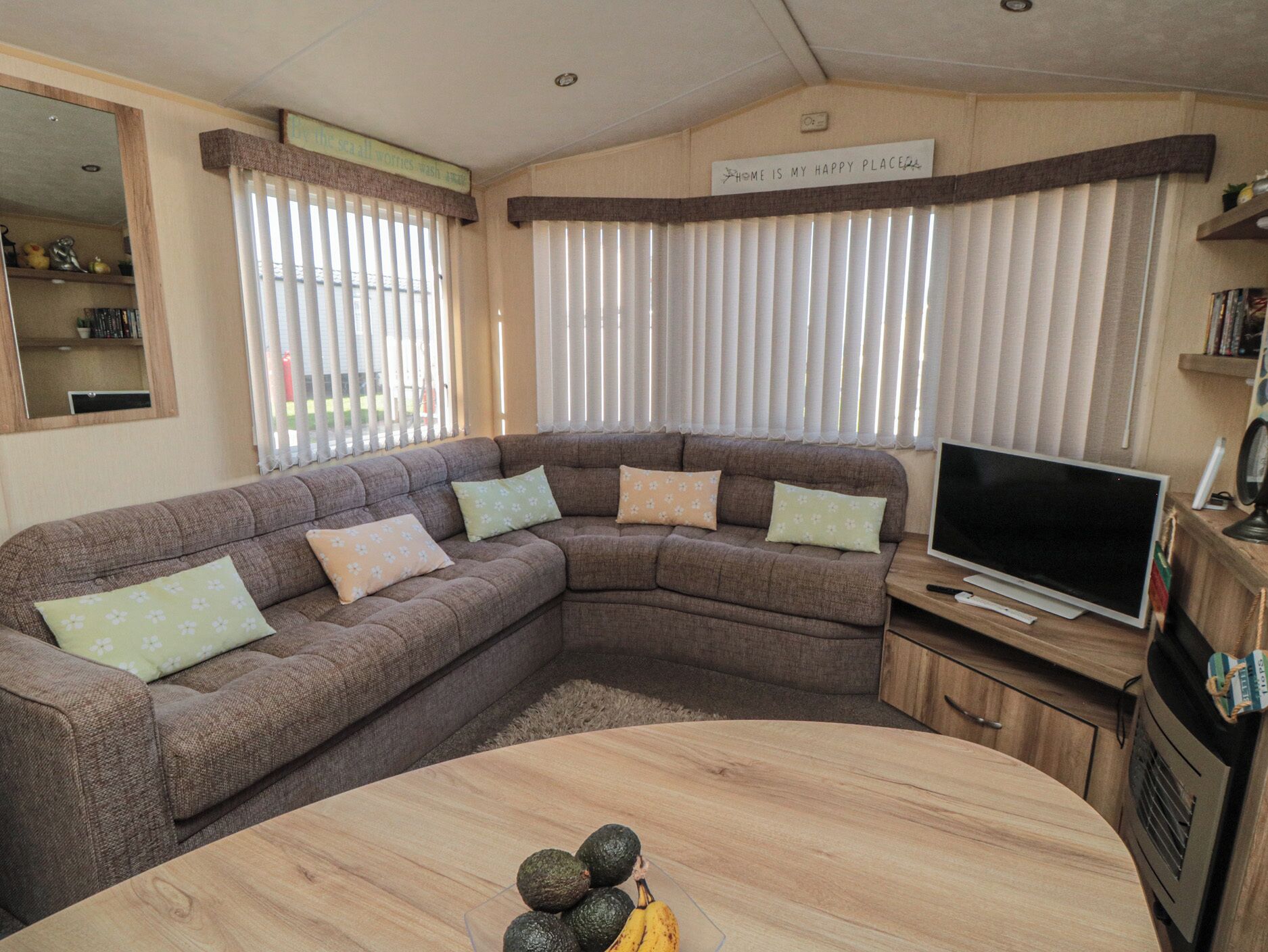 A living room with a sectional sofa, a flat-screen TV, a wooden coffee table, and a sign above the windows.