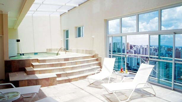 A modern indoor pool area with a city view, a hot tub, and a staircase leading to an upper level. - HOTEL PERDIZES - Flat Executivo - 1402 (São Paulo)