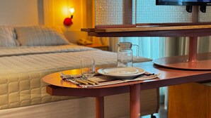 A neatly made bed with a quilted cover, a bedside table with a glass pitcher, a plate, and cutlery, and a television on a stand. - HOTEL PERDIZES - Flat Executivo - 1402 (São Paulo)