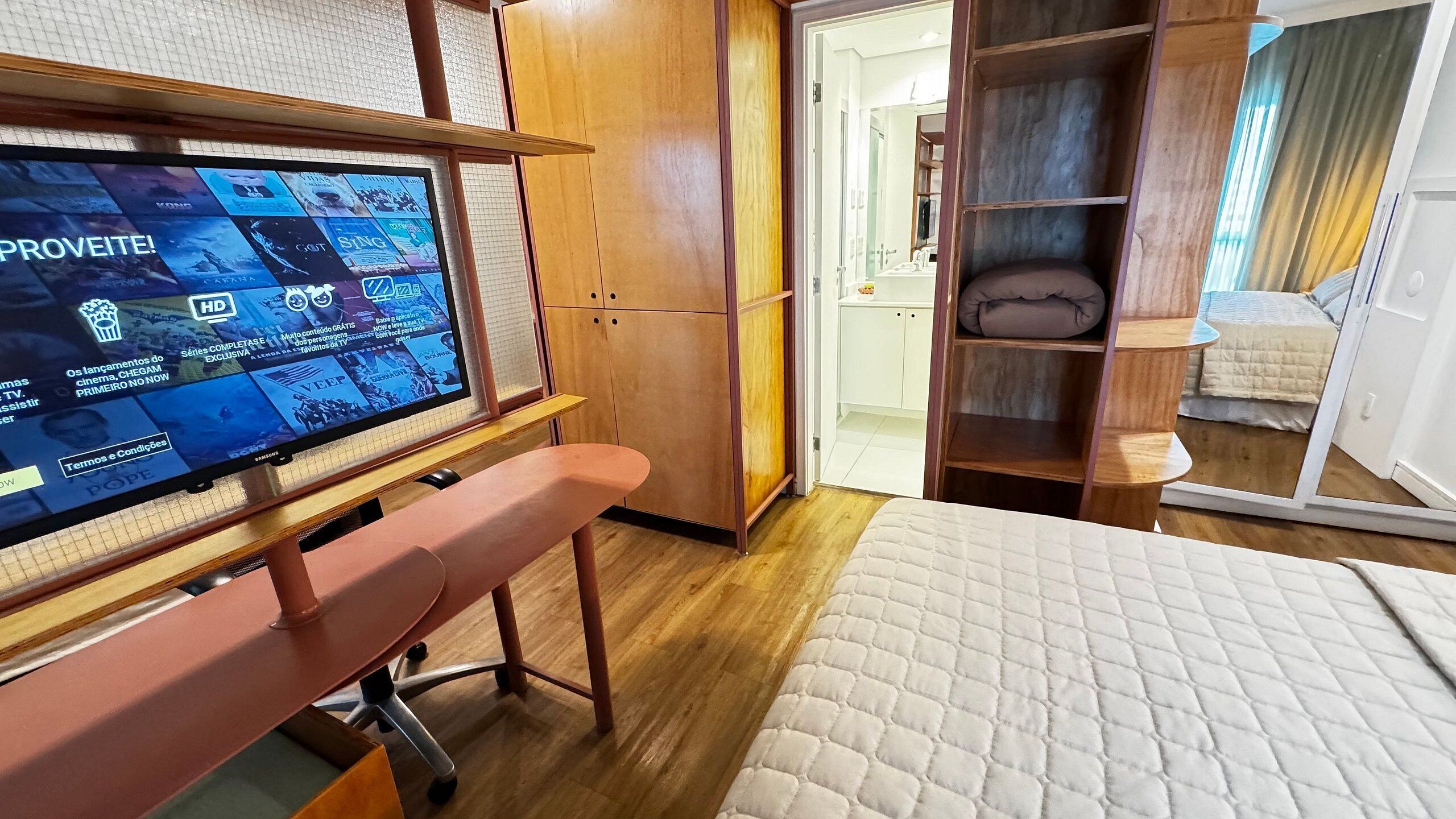Quarto de hotel com televisão exibindo um guia de programação, armário de madeira, um banco, cama com cobertura acolchoada e um espelho refletindo o ambiente.