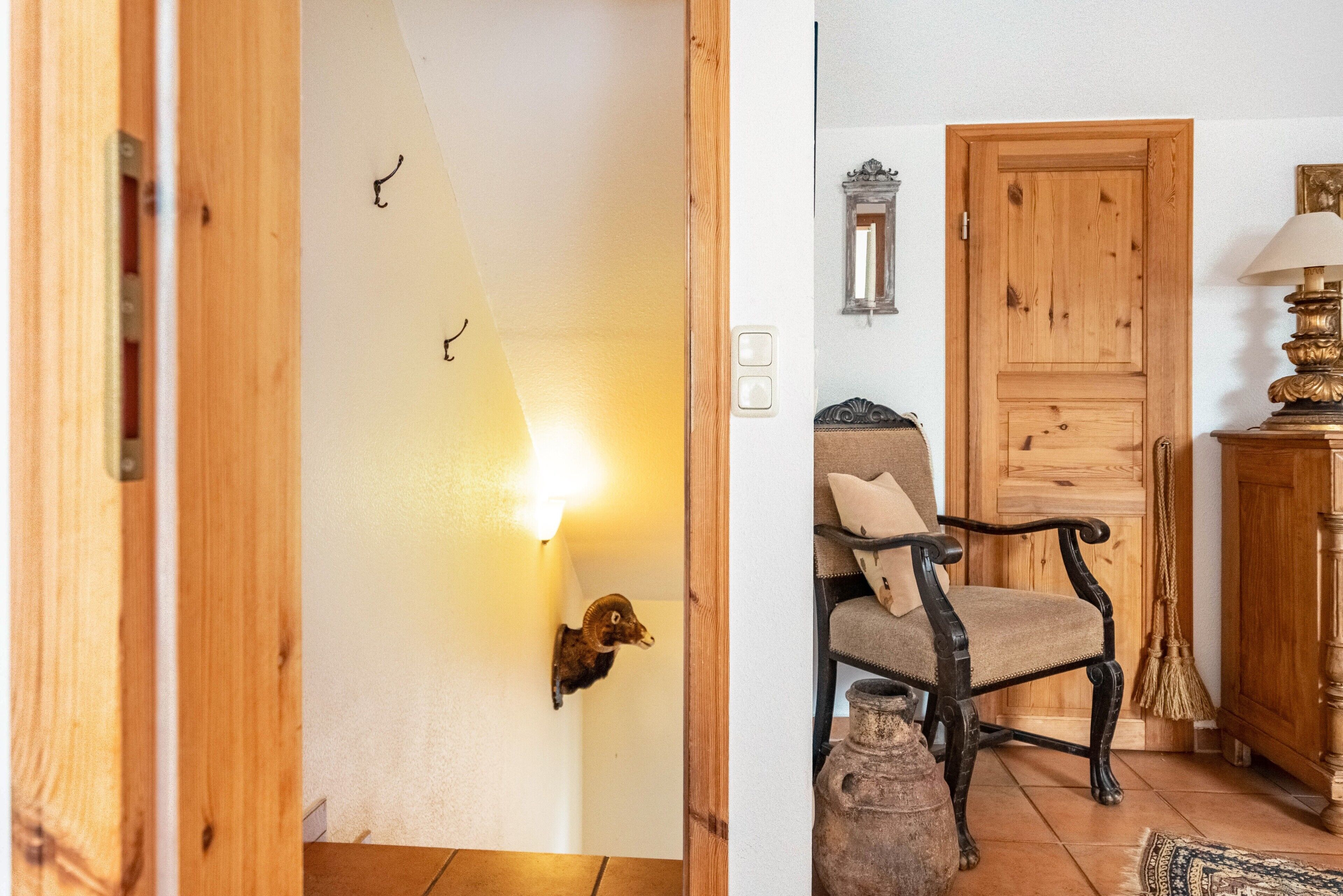 Eine Holztür, eine Wandleuchte, ein Sessel mit Kissen und ein Blumenvasen auf dem Boden.