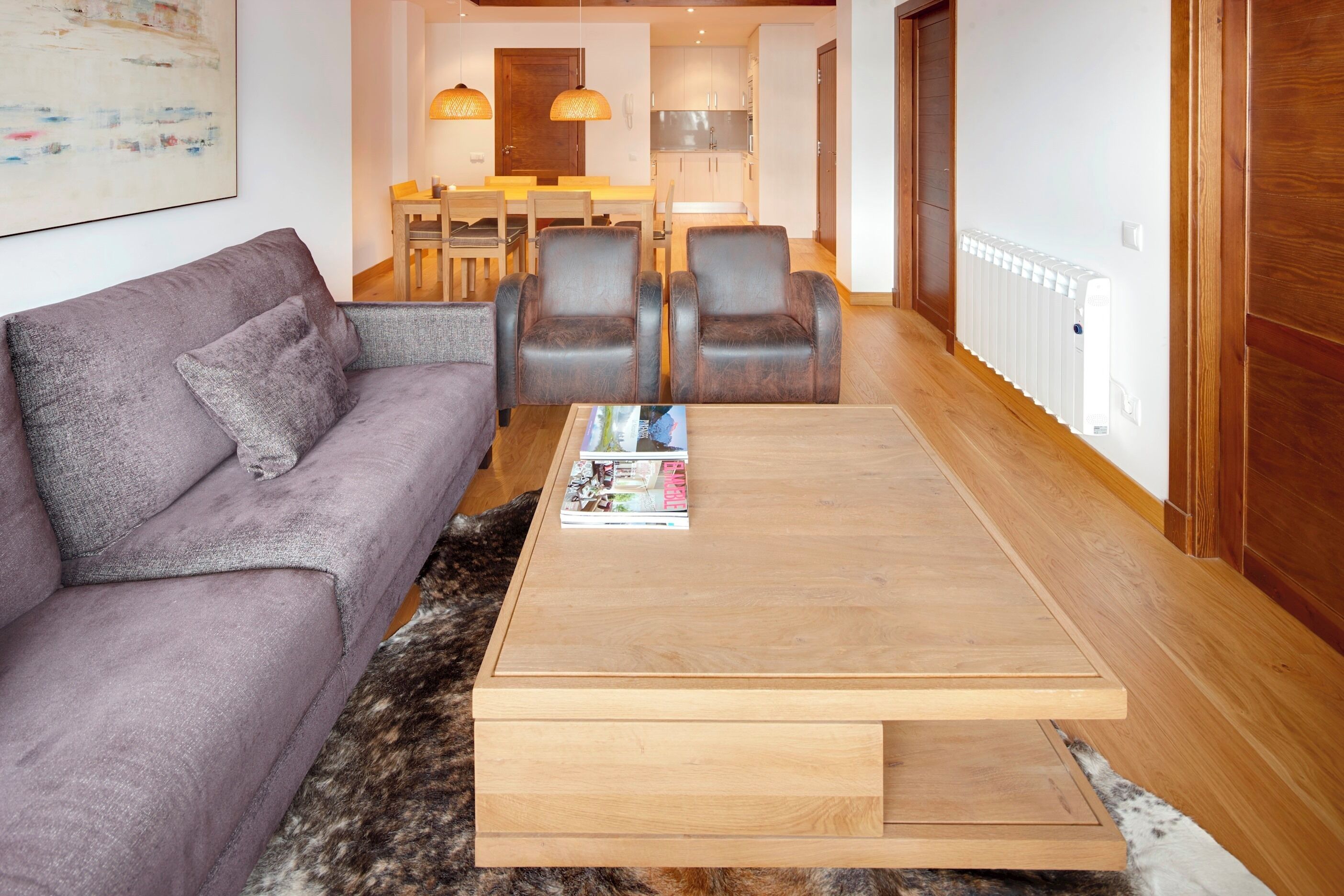 A modern living room with a sofa, two armchairs, a coffee table, and a rug. There is a dining area with a table and chairs in the background.