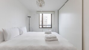 A minimalist bedroom with a large bed, a floor lamp, and a view of buildings through the window. - Modern King’s Cross Central Apartment (London)