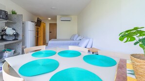 A compact living space with a kitchenette, a dining area with a white table and turquoise placemats, a sofa bed with pillows, and a potted plant. - Cupe Suites #118 Beachfront by Carpediem (Ipojuca)