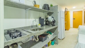 A kitchen with granite countertops, a gas stove, a sink, and a refrigerator. There are shelves above the counter with various items, including a coffee maker and a basket. - Cupe Suites #118 Beachfront by Carpediem (Ipojuca)