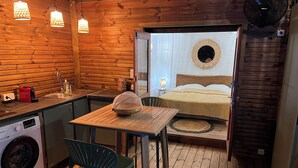 A compact room with a wooden interior, a bed with a circular mirror above it, a small table with a chair, a washing machine, and a fan.