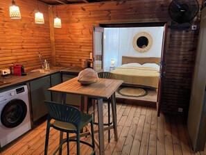 A compact room with a wooden interior, a bed with a circular mirror above it, a small table with a chair, a washing machine, and a fan. - "TI-NAGE " Unusual beaches 5 min walk (Sainte-Anne)