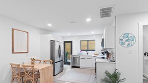 A modern kitchen with white cabinetry, a stainless steel refrigerator, and a dining area with a wooden table and chairs. - Small Serenity 1•Fully Remodeled•Beach Gear•Bikes• (Miramar Beach)