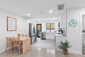 A modern kitchen with white cabinetry, a stainless steel refrigerator, and a dining area with a wooden table and chairs. - Small Serenity 1•Fully Remodeled•Beach Gear•Bikes• (Miramar Beach)