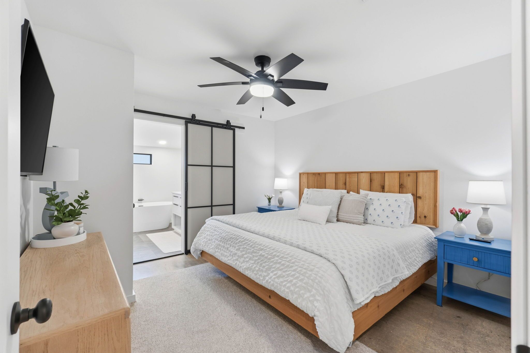 Un dormitorio con cama, ventilador de techo, cabecera de madera, mesitas de noche con lámparas y una puerta corrediza que da a otra habitación.