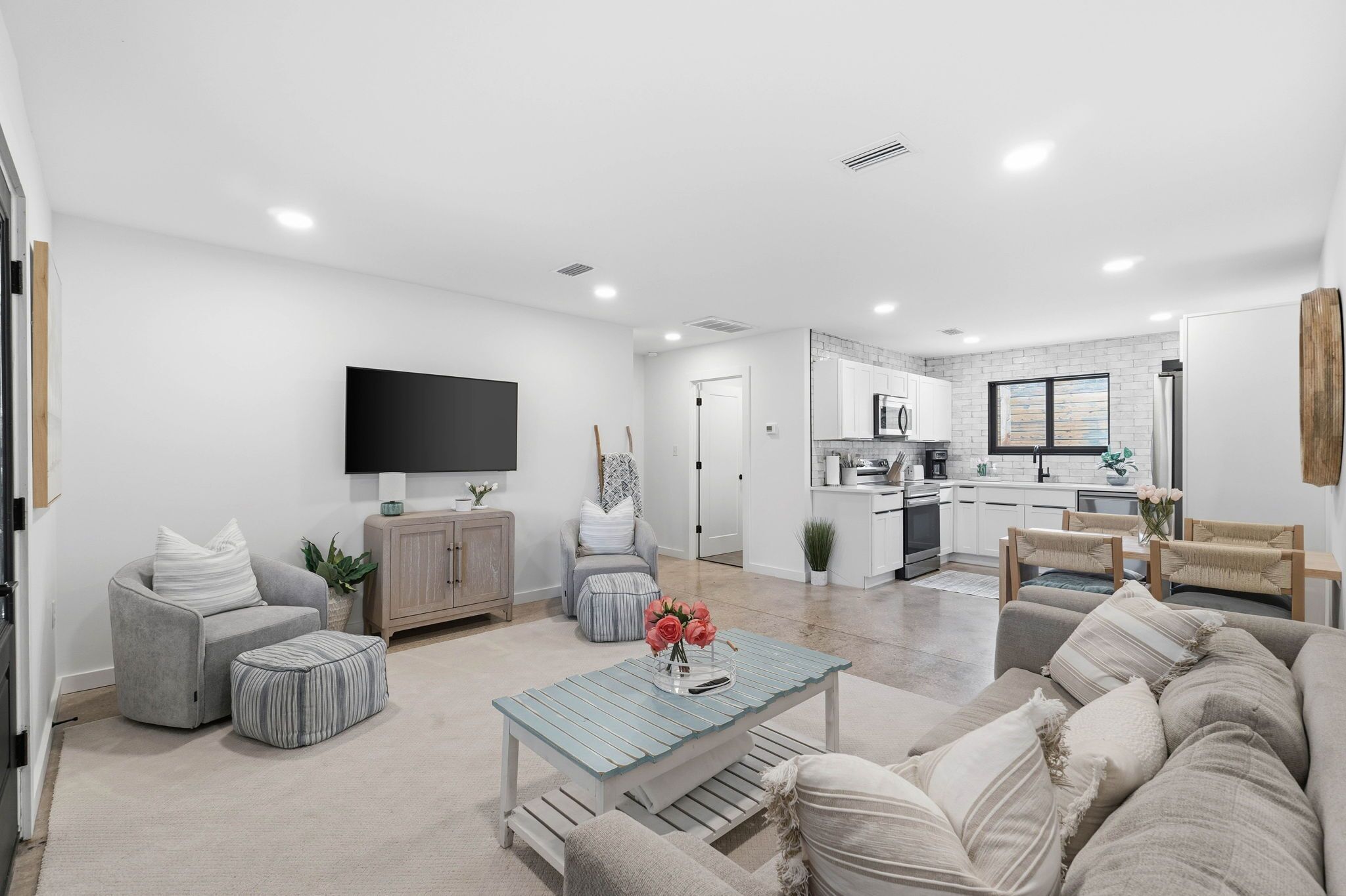 A modern living room with a sofa, ottoman, and coffee table, a flat-screen TV, and a kitchen in the background.