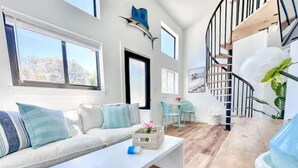 Uma sala de estar moderna com um sofá branco, uma mesa de centro e piso de madeira. Há uma escada com corrimão preto e vista para o exterior através de grandes janelas.