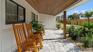 A covered outdoor seating area with wooden chairs and potted plants, a stone-paved patio, and a view of a residential area with palm trees. - Small Serenity 4•Fully Remodeled•Beach Gear•Bikes (Miramar Beach)