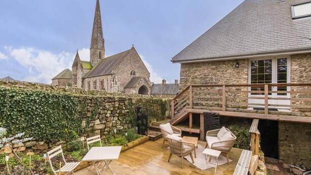 En bygning i sten med en træterrasse, udendørs siddepladser og en kirke i baggrunden.