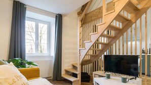 En moderne stue med en trætrappe, et stort fladskærms-tv, en hvid sofa og et vindue med gardiner.