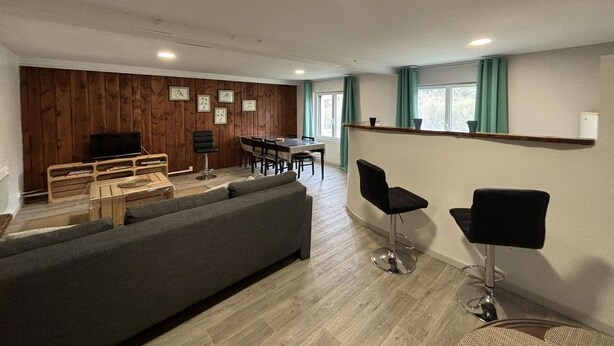 A modern living room with a grey sofa, wooden panel wall, dining table with chairs, and two black bar stools. - Modern apartment with country view - 2 guests (Le Vernet)