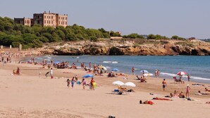 A beach with people sunbathing and swimming, a large building in the background, and rocky cliffs. - Bright apartment with terrace and parking near the beach (Tarragona)