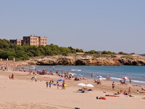 Ein Strand mit badenden und sonnenden Personen, ein großes Gebäude im Hintergrund und felsige Klippen.
