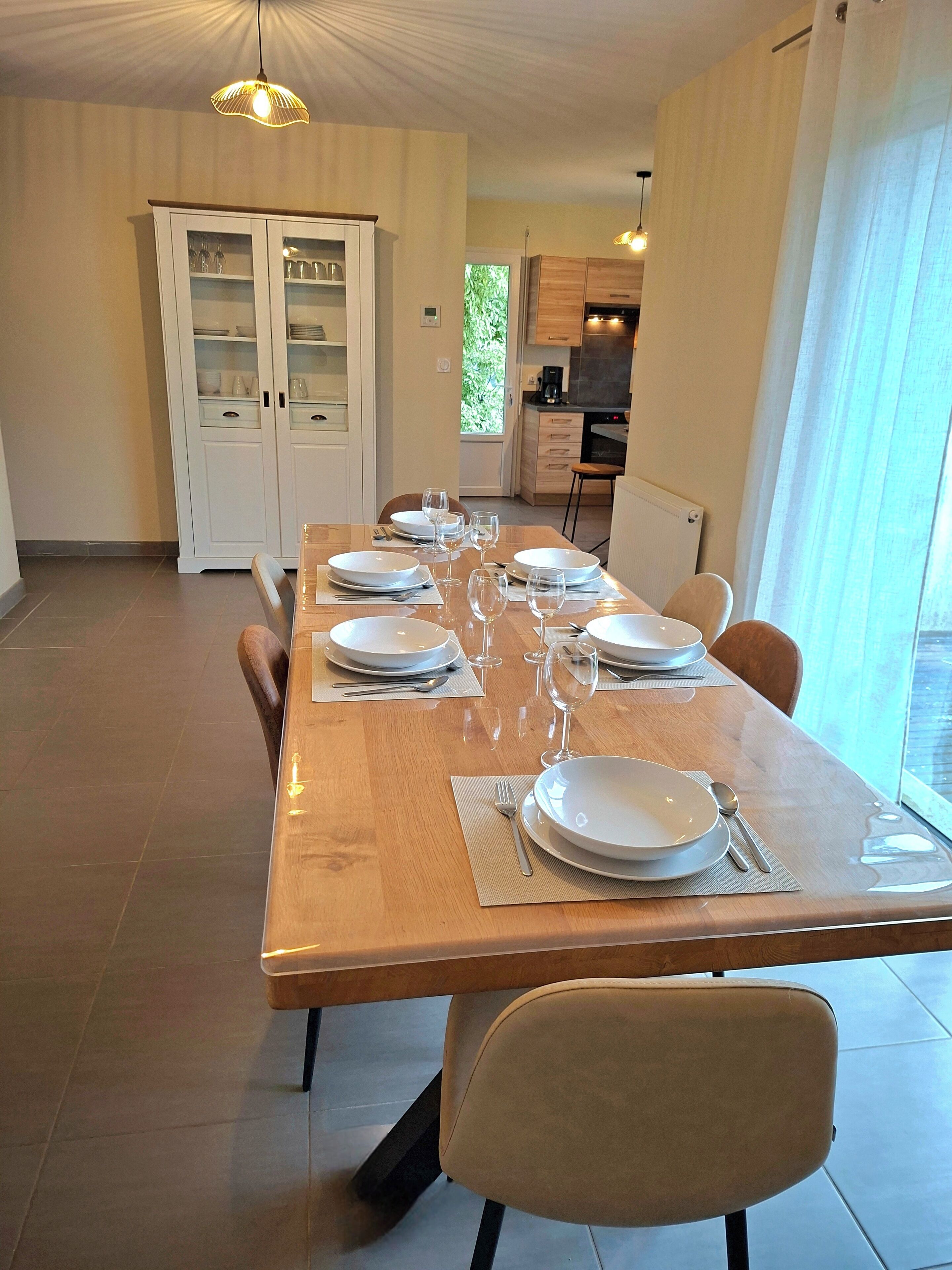 Un'area pranzo con un lungo tavolo di legno apparecchiato per sei persone, circondato da sedie. Si nota un armadietto bianco con ante in vetro sullo sfondo.
