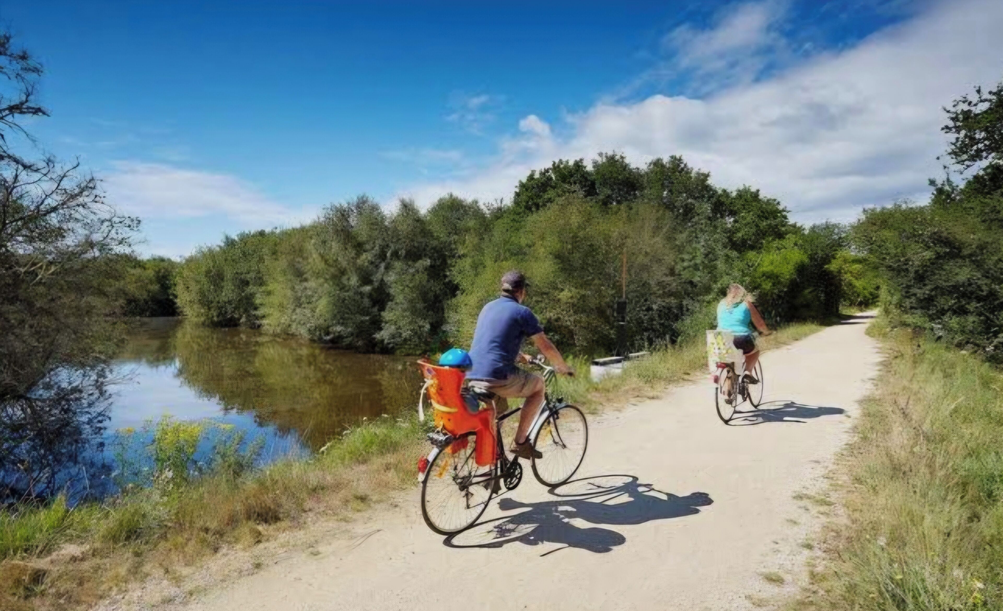 Two people cycling on a dirt path beside a calm river, surrounded by dense greenery.