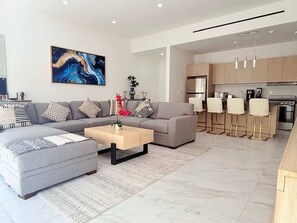 A modern living room with a grey sofa, a wooden coffee table, and a kitchen area with bar stools. - 2BR Downtown Cabo Walk to Beach & Dining (Cabo San Lucas)