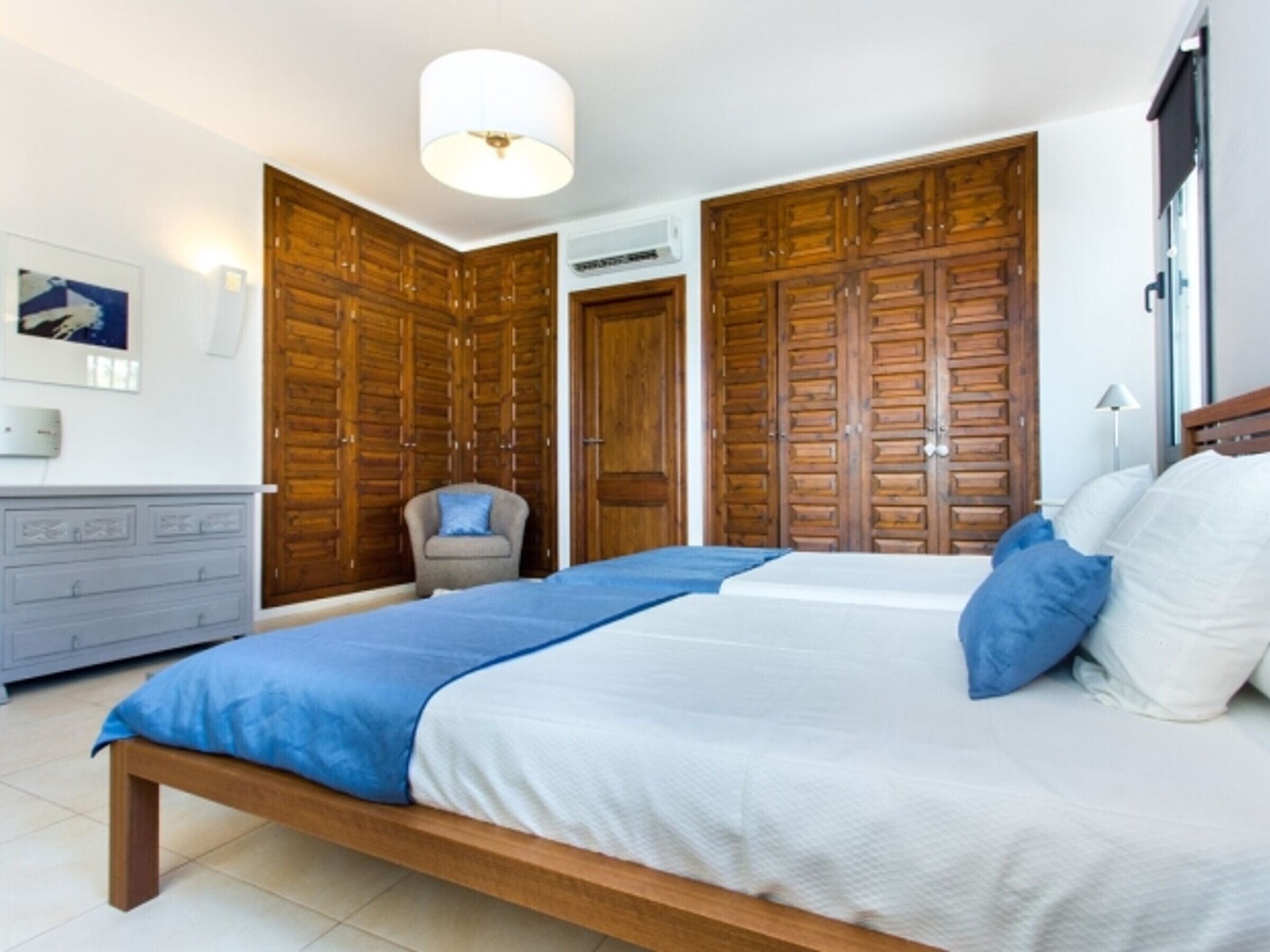 Un dormitorio con una cama grande, armarios de madera, un aparador gris y una lámpara en el techo.