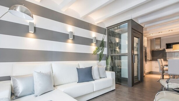 Een moderne woonkamer met een witte bank, een glazen deur naar een keuken, en een plafond met inbouwspots.