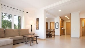 Ein geräumiges Wohnzimmer mit einer Couch, einem Couchtisch und einem Essbereich mit Stühlen und Tisch. Auf dem Couchtisch steht eine Lampe und an der Wand hängt ein Gemälde. Das Zimmer hat große Fenster und blickt auf Bäume hinaus.