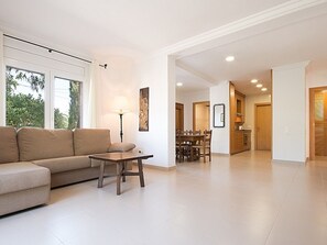 A spacious living room with a sofa, a coffee table, and a dining area with chairs and a table. There is a lamp on the coffee table and a painting on the wall. The room has large windows and a view of trees outside. - Renovated Villa in Salou: 4 bdrm, 4 bath, barbecues, near beach (Salou)