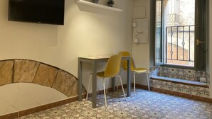 Ein Zimmer mit an der Wand montiertem Fernseher, einem kleinen Tisch mit zwei gelben Stühlen, einer Bank und einem gemusterten Boden.