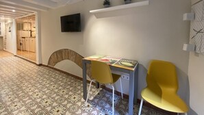 Ein Zimmer mit einem gemusterten Boden, einem Schreibtisch mit Stuhl, einem Fernseher an der Wand und einem Regal mit kleinen Pflanzen.