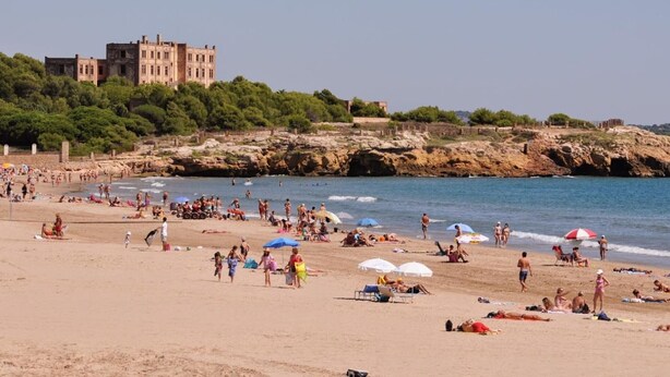 A beach with people sunbathing and swimming, a large building in the background, and rocky cliffs. - Bright apartment in the historic center of Tarragona (Tarragona)