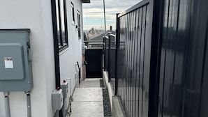 A narrow pathway leading to a gated entrance, flanked by a building with external electrical boxes and a metal fence. - BRAND NEW MODERN 3BR LANEWAY HOUSE (Vancouver)