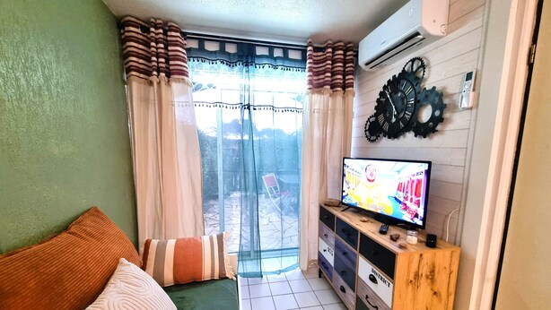 Ein Wohnzimmer mit einem grünen Sofa, einem Fernseher auf einem Holzschrank und einer Glastür, die zu einem Außenbereich führt.