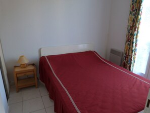 Une chambre à coucher avec un lit, une table de chevet avec une lampe, une fenêtre avec des rideaux et un radiateur.