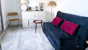 Un espacio habitable compacto con un sofá azul oscuro, dos cojines rojos, una pequeña mesa redonda, un sillón de mimbre, un armario blanco y dos lámparas.