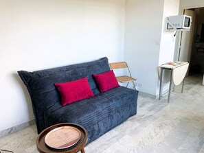 A dark blue sofa with red cushions, a wicker chair, a small table with a magazine, and a white microwave on a white cabinet. - Cozy studio, AC and parking - 2 guests in La Grande-Motte (La Grande-Motte)