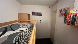Et lille, smalt værelse med en enkelt seng, et træskab, et billede i ramme på væggen og en reol med bøger.