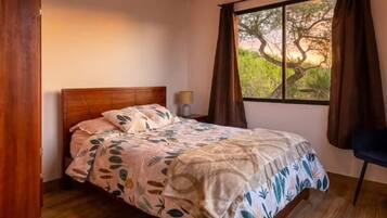 A bedroom with a bed, a nightstand, a lamp, a window with curtains, and a view of trees.