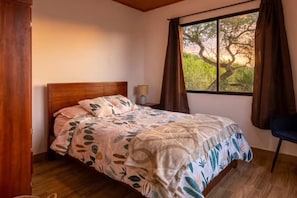 A bedroom with a bed, a nightstand, a lamp, a window with curtains, and a view of trees. - Modern Country House with Heated Pool in Yunguilla Valley Near Cuenca Ecuador (Santa Isabel)
