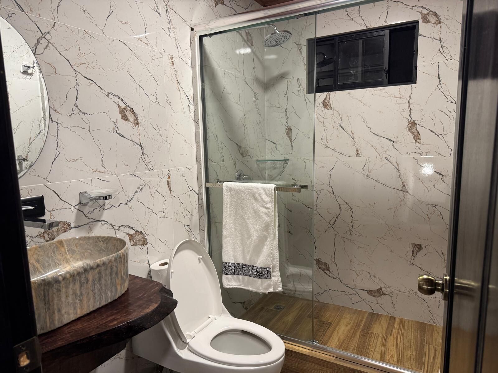 Ein Badezimmer mit Marmorfliesenwand, einer Glaskabinedusche, Holzboden und einer weißen Toilette.