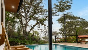 Un espace piscine avec des chaises longues en bois, une piscine à l’eau d’un bleu limpide et des arbres tout autour.