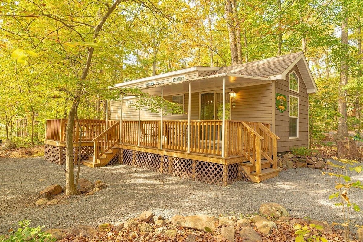 A cabin with a wooden deck, a covered porch, and a gravel driveway surrounded by trees.