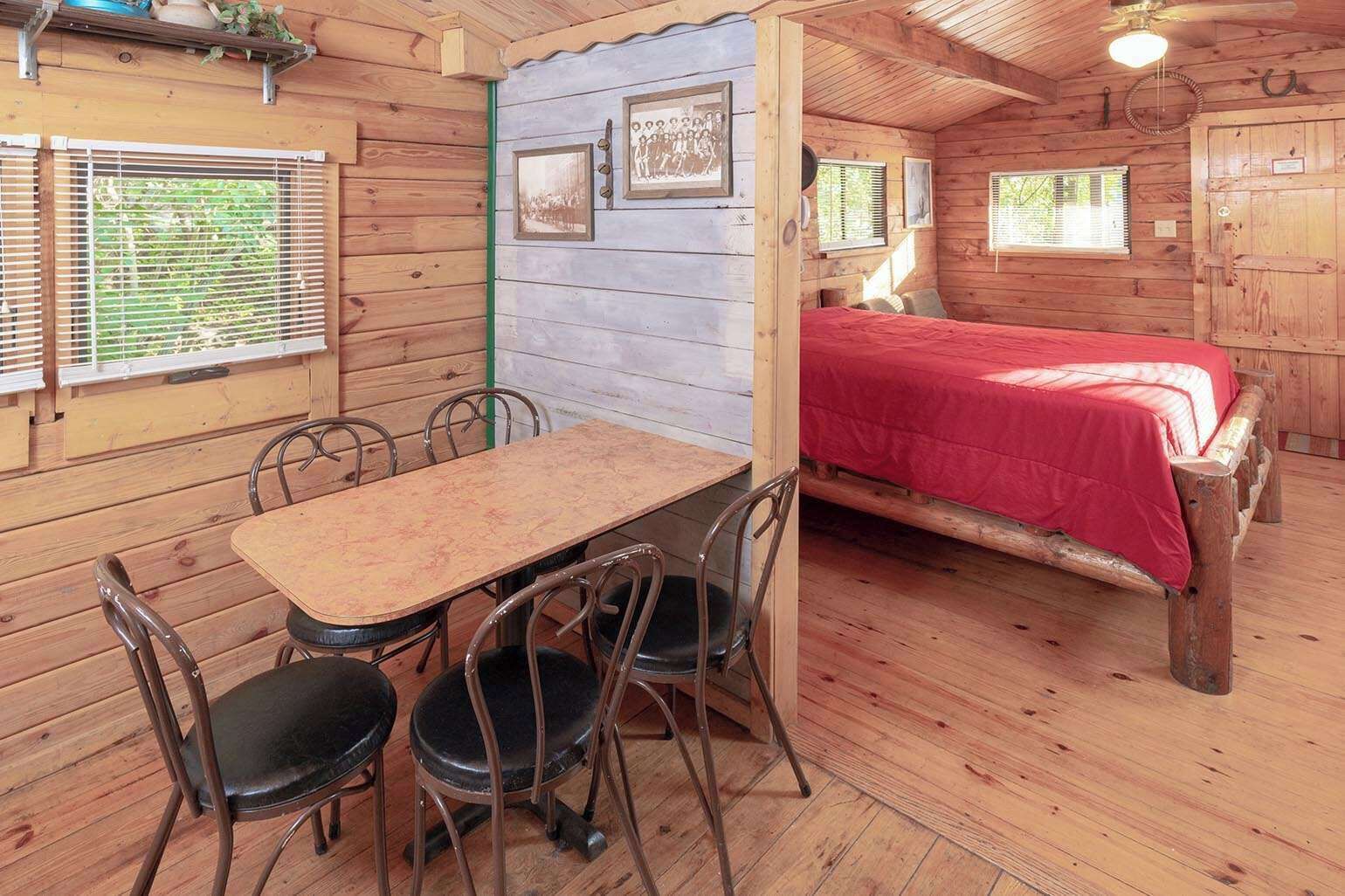 A wooden cabin interior with a dining table, four chairs, a bed with a red cover, and a small window.