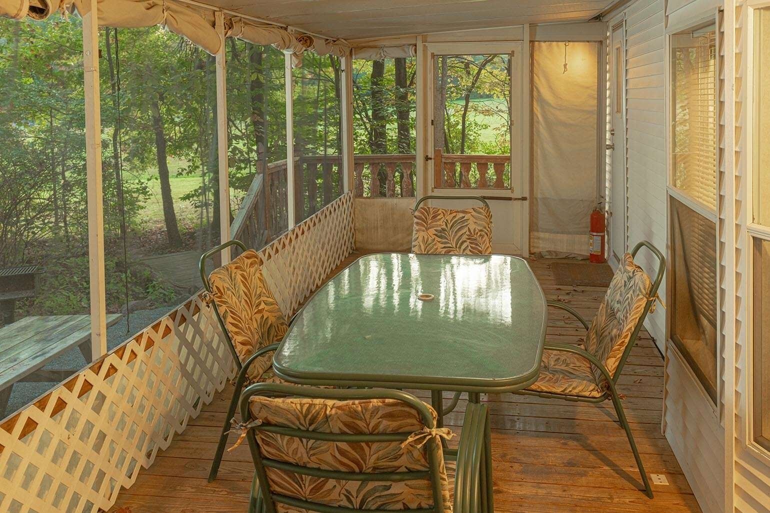 Un porche cubierto con una mesa y sillas, una terraza de madera y vistas a los árboles circundantes.