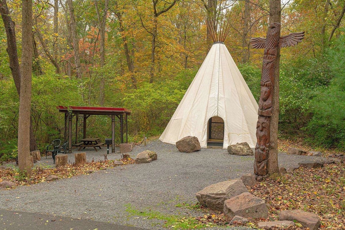 A teepee with a wooden door, a picnic table, and a covered bench area in a forest setting.