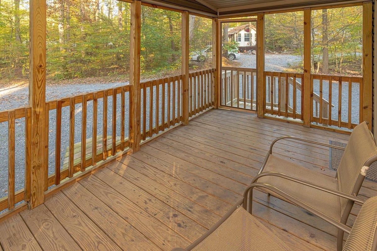 A covered wooden deck with a bench, surrounded by a wooden railing, overlooking a gravel driveway and a house.