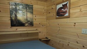 Puinen mökkihuone, jossa on sänky, metsäpolkua esittävä maalaus ja kehystetty kuva kissasta.