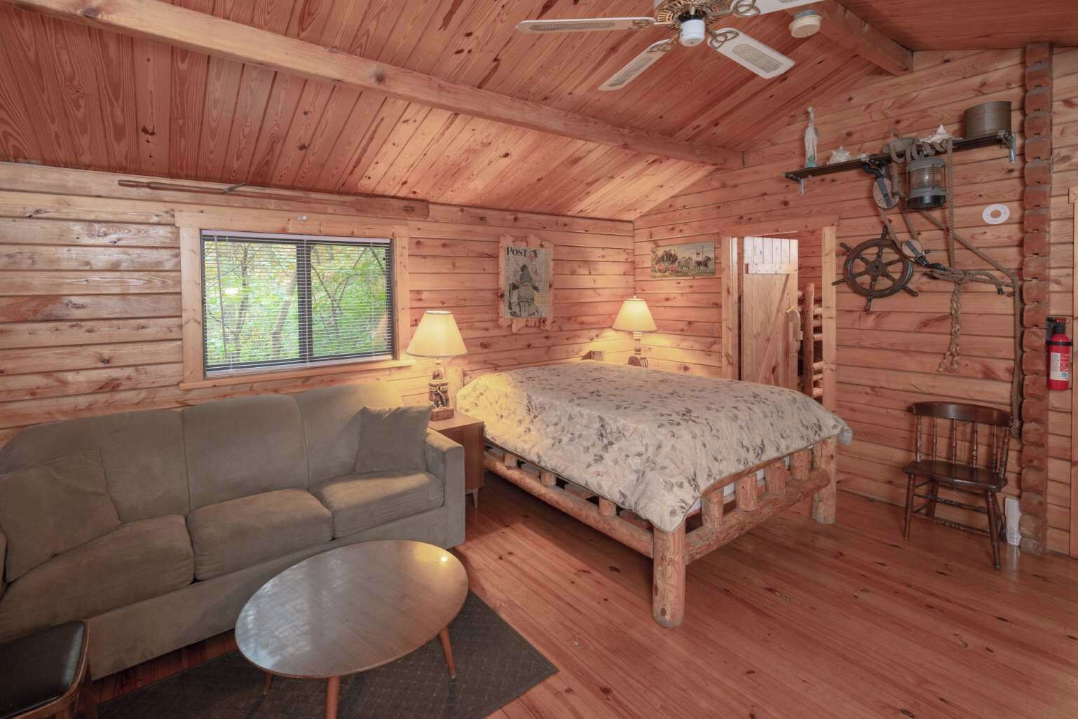 A log cabin room with a bed, a sofa, a small table, a ceiling fan, and a window with blinds.