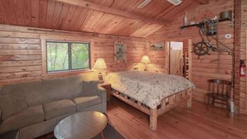 A log cabin room with a bed, a sofa, a small table, a ceiling fan, and a window with blinds.